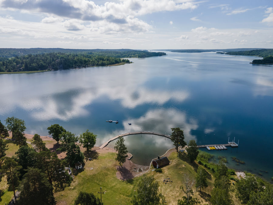 Flygfoto över en badplats vid en stor sjö, omgiven av grönskande skog, med brygga och båtar vid vattnet.