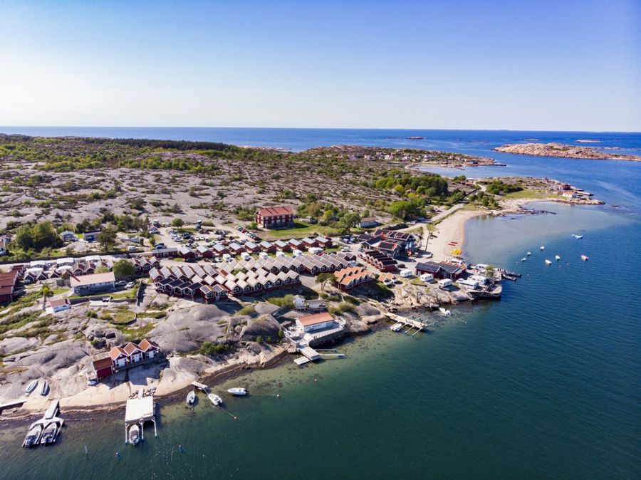Flygfoto över Ramsvik Stugby & Camping med sina röda stugor, campingtomter och badstrand omgiven av hav och bohuslänska klippor.